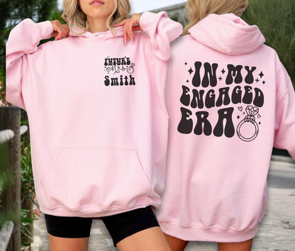 May include: A light pink hooded sweatshirt featuring black text and graphics. The front displays "Future Mrs. Smith" with a ring icon. The back features the text "IN MY ENGAGED ERA" in a bold, wavy font, accompanied by small star and diamond ring icons.