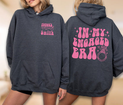 May include: A dark charcoal gray hooded sweatshirt featuring pink text and graphics. The front displays "FUTURE MRS SMITH" with a ring icon. The back features the large pink text "IN MY ENGAGED ERA" accompanied by star, heart, and diamond ring graphics.