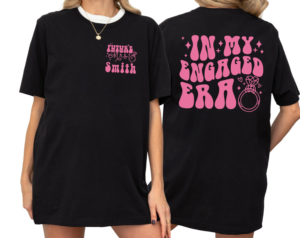 May include: A black t-shirt featuring pink text and graphics. The front left chest displays 'Future Mrs. Smith' with a small ring icon. The back features large, wavy pink text reading 'IN MY ENGAGED ERA' alongside a diamond ring graphic and stars.