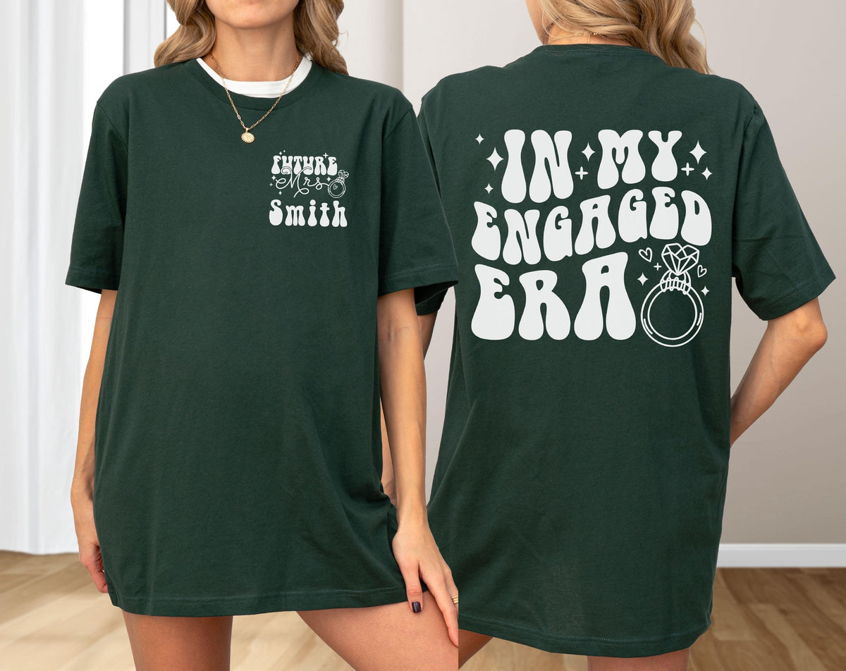 May include: A dark green t-shirt featuring white graphic text. The front left chest displays the text "Future Mrs. Smith" with a small ring icon. The back features the large white text "IN MY ENGAGED ERA" accompanied by a diamond ring illustration.