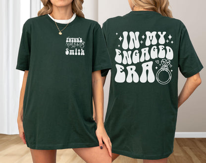 May include: A dark green t-shirt featuring white graphic text. The front left chest displays the text "Future Mrs. Smith" with a small ring icon. The back features the large white text "IN MY ENGAGED ERA" accompanied by a diamond ring illustration.