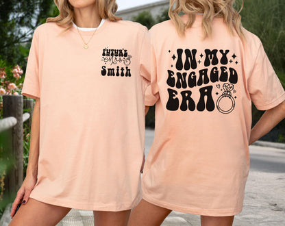 May include: A peach-colored t-shirt featuring black text and a ring graphic. The front left chest displays 'Future Mrs. Smith' with a small ring. The back features large, bold, wavy black text reading 'IN MY ENGAGED ERA' alongside a diamond ring icon.