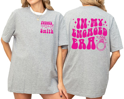 May include: A heather gray t-shirt featuring hot pink text and graphics. The front left chest displays the text "FUTURE MRS. SMITH" with a ring icon. The back features the large text "IN MY ENGAGED ERA" with a diamond ring and star accents.
