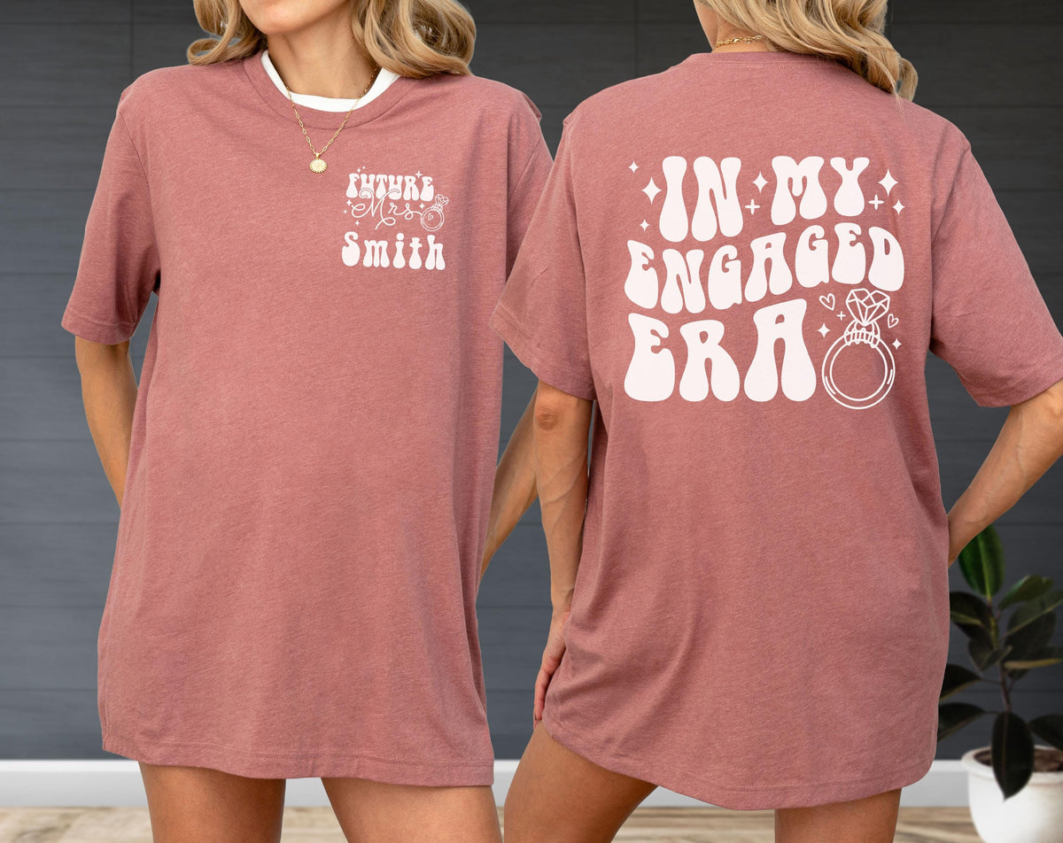 May include: A dusty rose t-shirt featuring white screen-printed text. The front left chest displays the text "FUTURE MRS SMITH" with a small ring icon. The back features the large text "IN MY ENGAGED ERA" accompanied by a diamond ring graphic.
