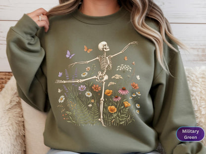 May include: A military green crewneck sweatshirt featuring a graphic print of a skeleton in a dancing pose surrounded by colorful wildflowers, ferns, and small butterflies. The design is centered on the chest of the long-sleeved pullover garment.