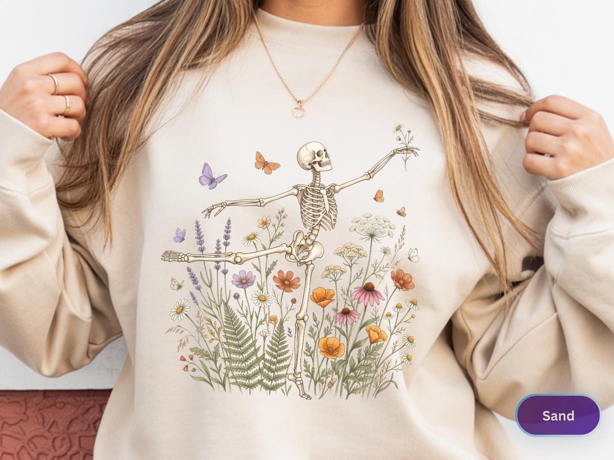 May include: A sand-colored crewneck sweatshirt featuring a graphic print of a skeleton dancing amidst a field of colorful wildflowers, ferns, and butterflies. The skeleton is posed with one leg extended and arms reaching out toward small flying insects.