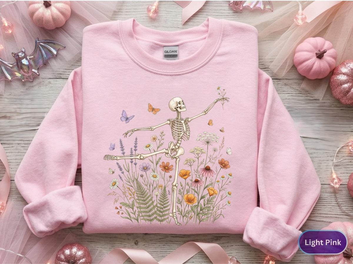May include: A light pink long-sleeve sweatshirt featuring a graphic print of a skeleton dancing amidst a field of colorful wildflowers, ferns, and small butterflies. The Gildan brand label is visible at the inner neckline of the garment.