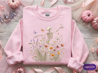 May include: A light pink long-sleeve sweatshirt featuring a graphic print of a skeleton dancing amidst a field of colorful wildflowers, ferns, and small butterflies. The Gildan brand label is visible at the inner neckline of the garment.