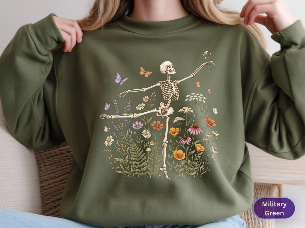 May include: A military green crewneck sweatshirt featuring a graphic print of a skeleton dancing amidst a field of colorful wildflowers and butterflies. The skeleton is depicted in a ballet pose, surrounded by daisies, poppies, and ferns.
