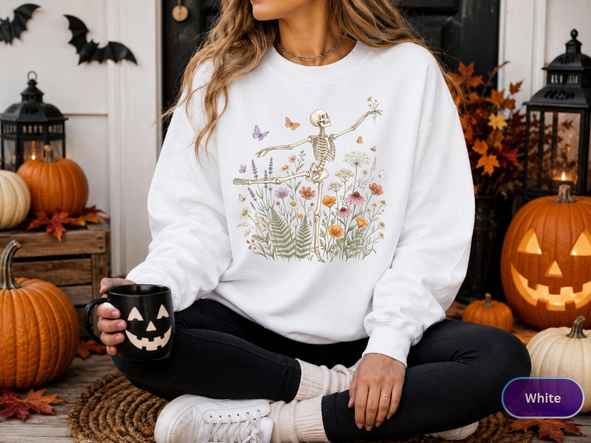 May include: A white long-sleeve sweatshirt featuring a graphic print of a skeleton dancing amidst a colorful field of wildflowers, ferns, and butterflies. The design includes various floral elements in shades of orange, purple, and green.