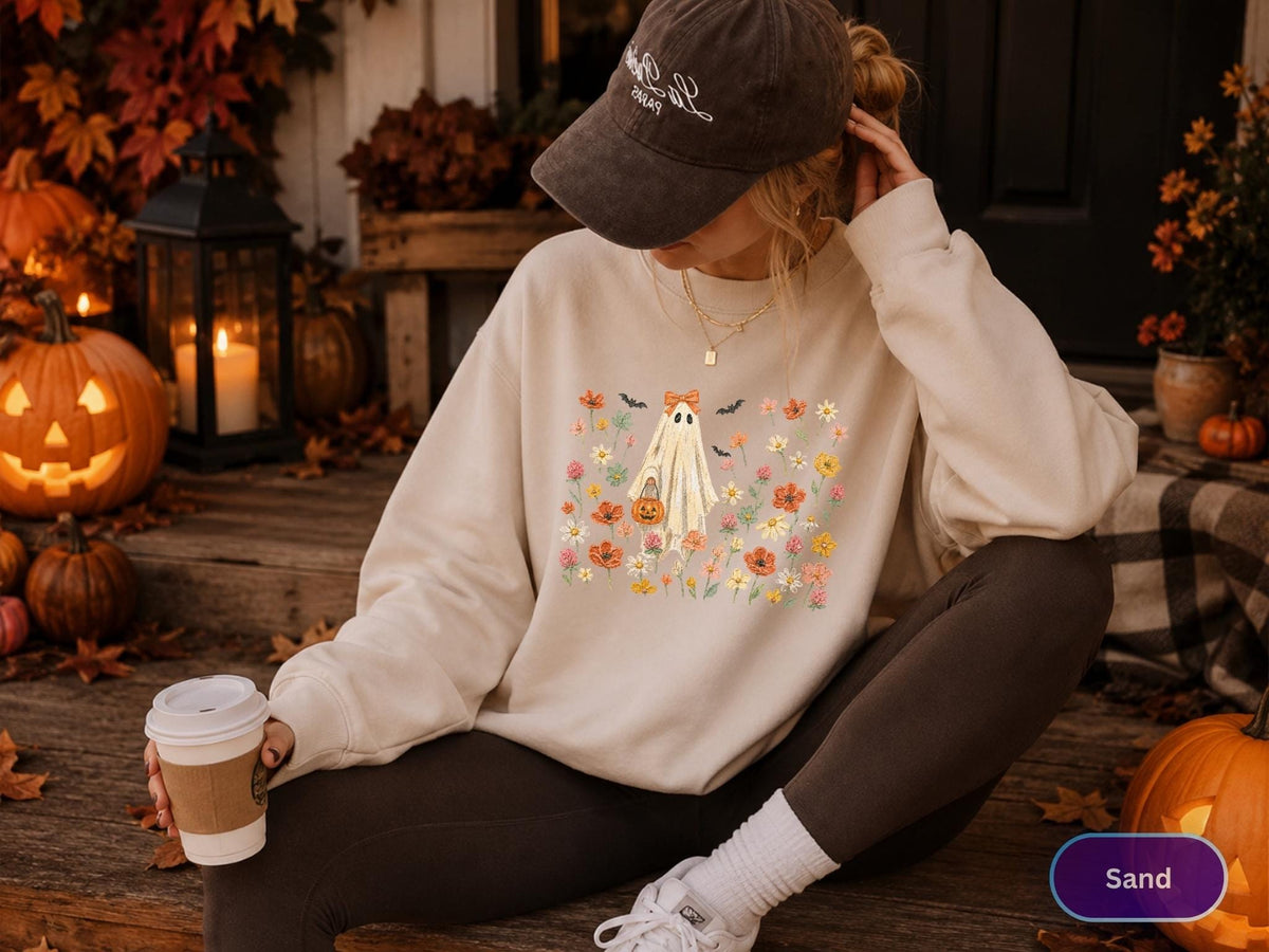 May include: A sand-colored crewneck sweatshirt featuring a central graphic of a ghost wearing a bow, holding a pumpkin bucket, and surrounded by colorful wildflowers and small bats. The sweatshirt has long sleeves and a relaxed fit.