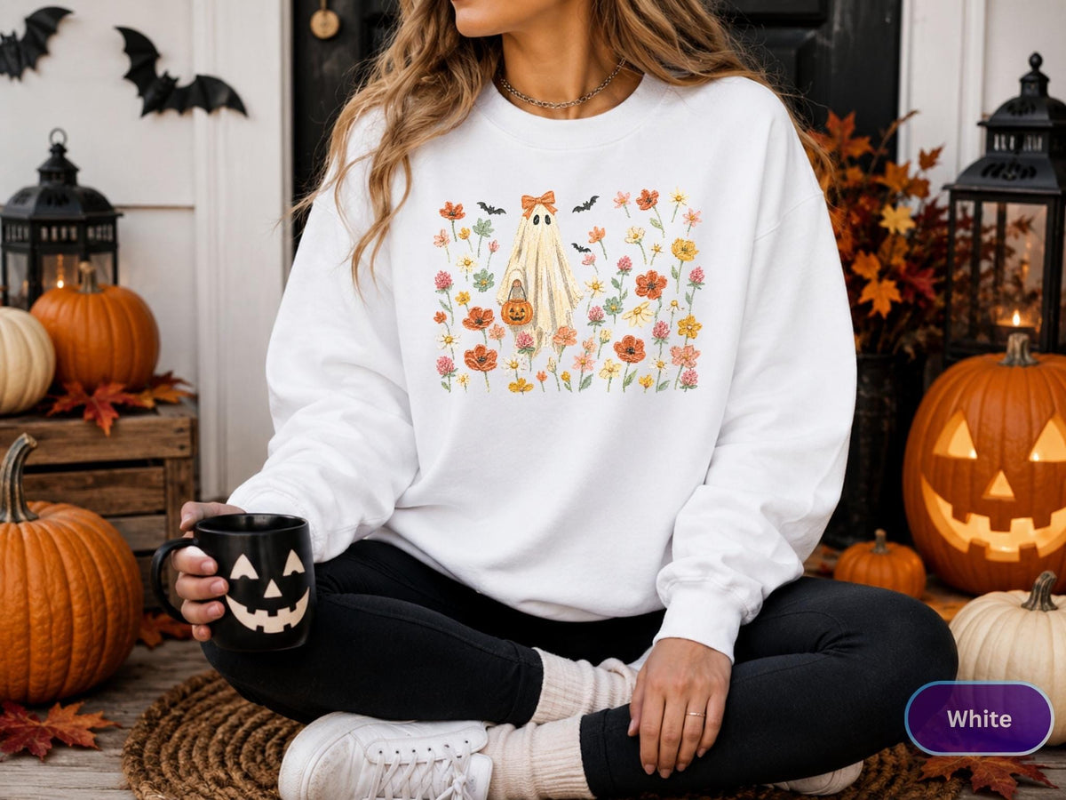 May include: A white crewneck sweatshirt featuring a central graphic of a ghost wearing an orange bow, holding a small pumpkin bucket, surrounded by a colorful field of wildflowers and small black bats in shades of orange, yellow, and pink.