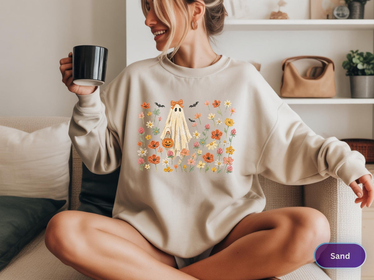 May include: A sand-colored crewneck sweatshirt featuring a central graphic of a ghost wearing an orange bow and holding a jack-o'-lantern bucket, surrounded by a colorful illustration of various wildflowers and small black bats.