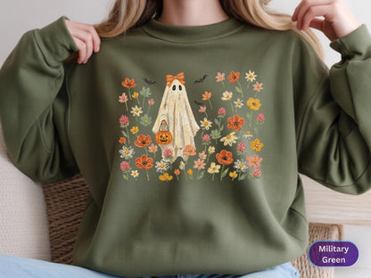 May include: A military green crewneck sweatshirt featuring a central graphic of a ghost wearing an orange bow and holding a jack-o'-lantern bucket. The ghost is surrounded by a colorful field of various orange, pink, and white wildflowers and bats.