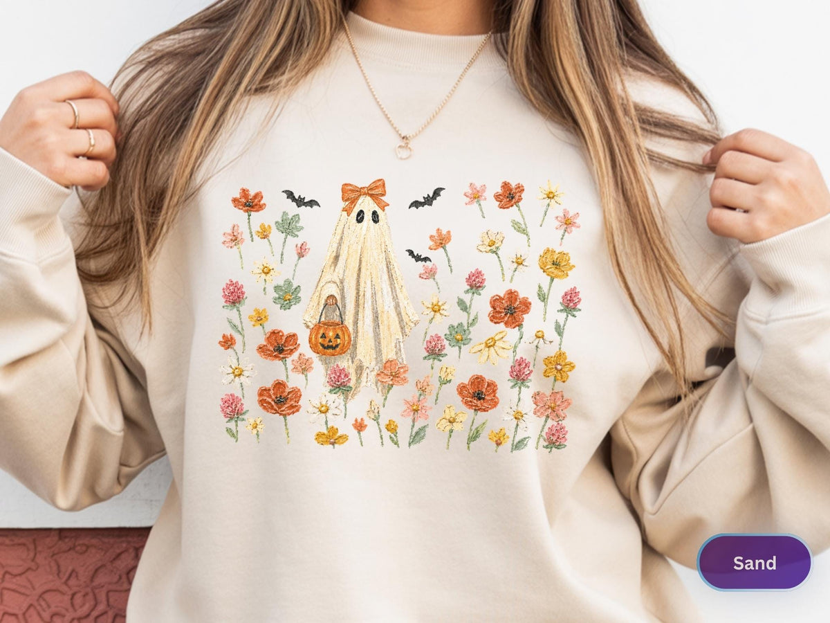 May include: A sand-colored crewneck sweatshirt featuring a central graphic of a ghost wearing an orange bow and holding a jack-o'-lantern bucket. The ghost is surrounded by various colorful wildflowers and small black bats in a field-like arrangement.
