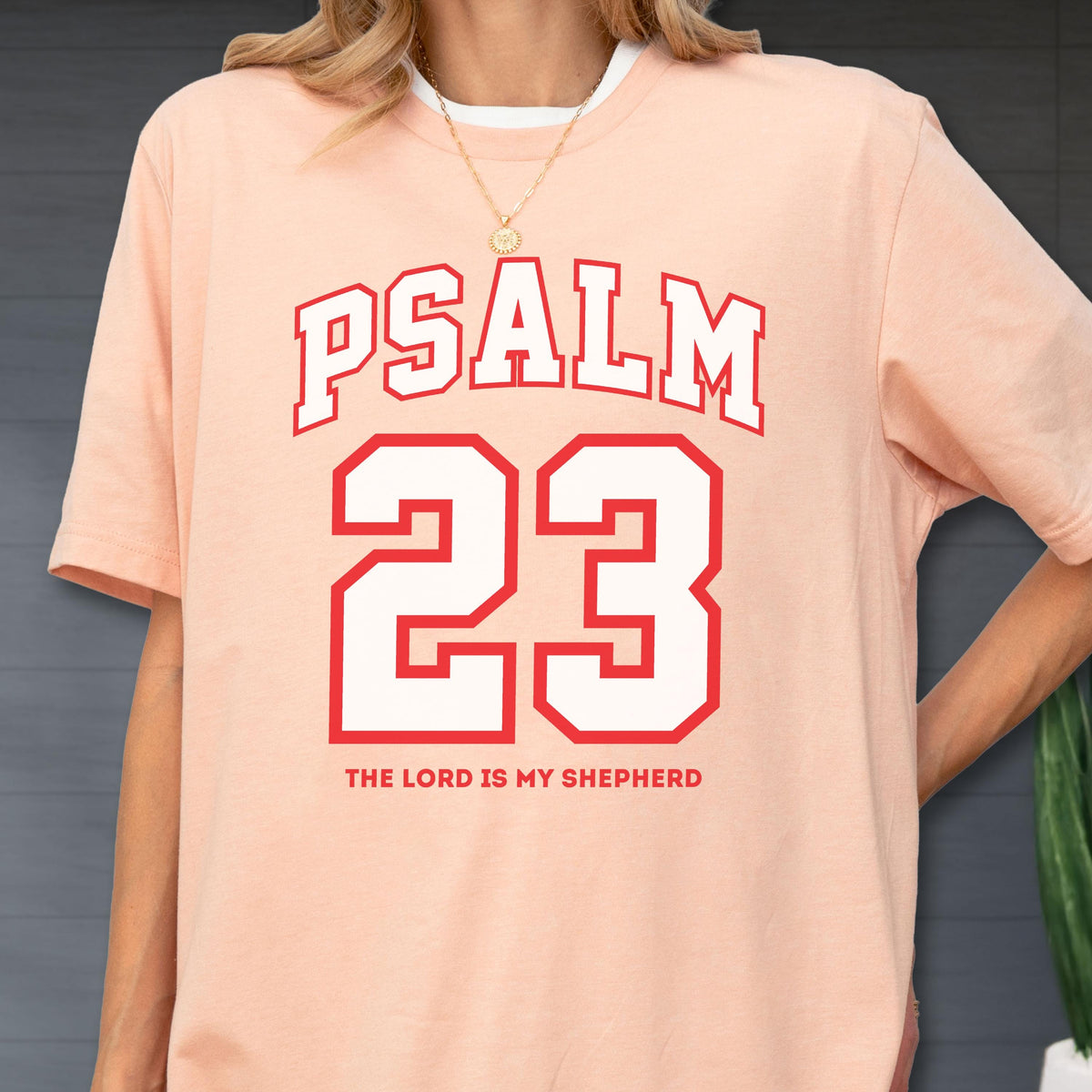 May include: A peach-colored short-sleeve t-shirt featuring the text PSALM 23 in a red varsity-style font, with the number 23 in large white block lettering outlined in red, and the phrase THE LORD IS MY SHEPHERD printed in red below the number.