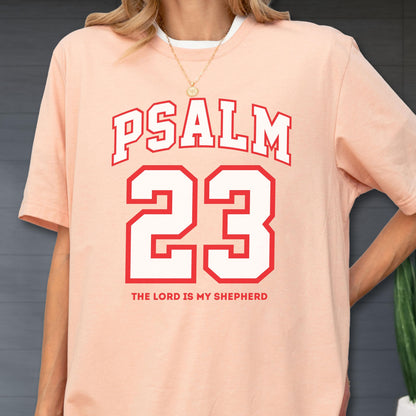 May include: A peach-colored short-sleeve t-shirt featuring the text PSALM 23 in a red varsity-style font, with the number 23 in large white block lettering outlined in red, and the phrase THE LORD IS MY SHEPHERD printed in red below the number.