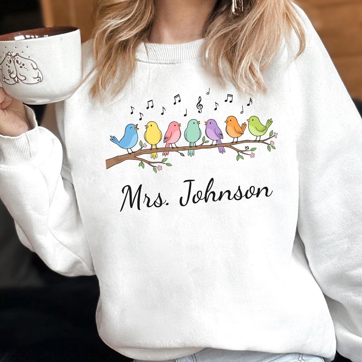 May include: A white crewneck sweatshirt featuring a graphic of seven colorful birds perched on a brown branch with musical notes above them. Below the branch, the text Mrs. Johnson is printed in a black cursive font on the front of the garment.