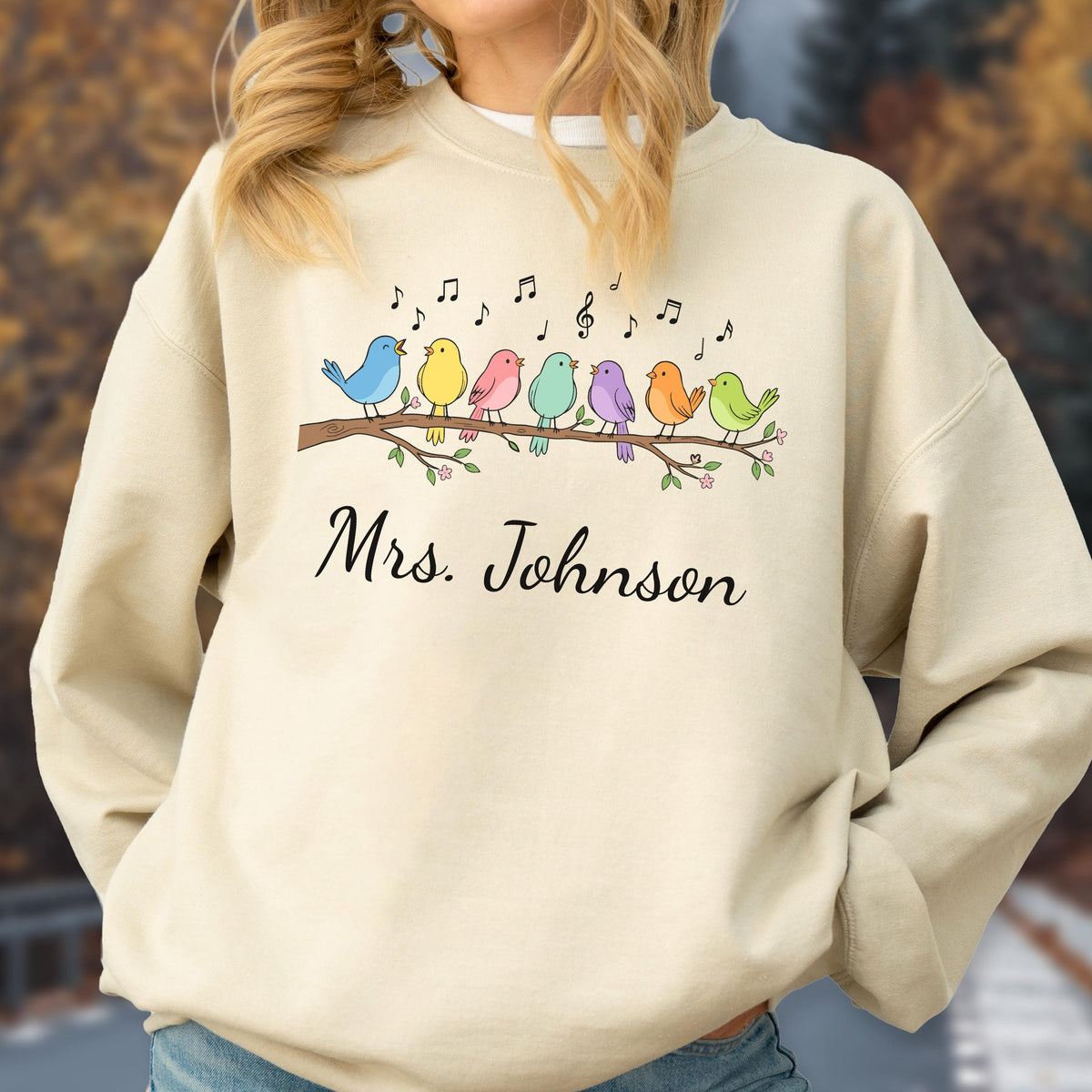 May include: A beige crewneck sweatshirt featuring a graphic of seven colorful birds perched on a brown branch with musical notes above them. The text "Mrs. Johnson" is printed in black cursive script below the bird illustration on the front.