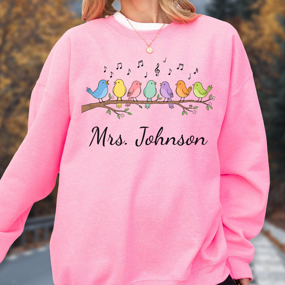 May include: A bright pink crewneck sweatshirt featuring a graphic of eight colorful birds perched on a brown tree branch with musical notes above them. The text "Mrs. Johnson" is printed in black cursive script below the bird illustration.