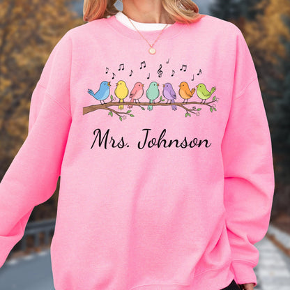 May include: A bright pink crewneck sweatshirt featuring a graphic of eight colorful birds perched on a brown tree branch with musical notes above them. The text "Mrs. Johnson" is printed in black cursive script below the bird illustration.
