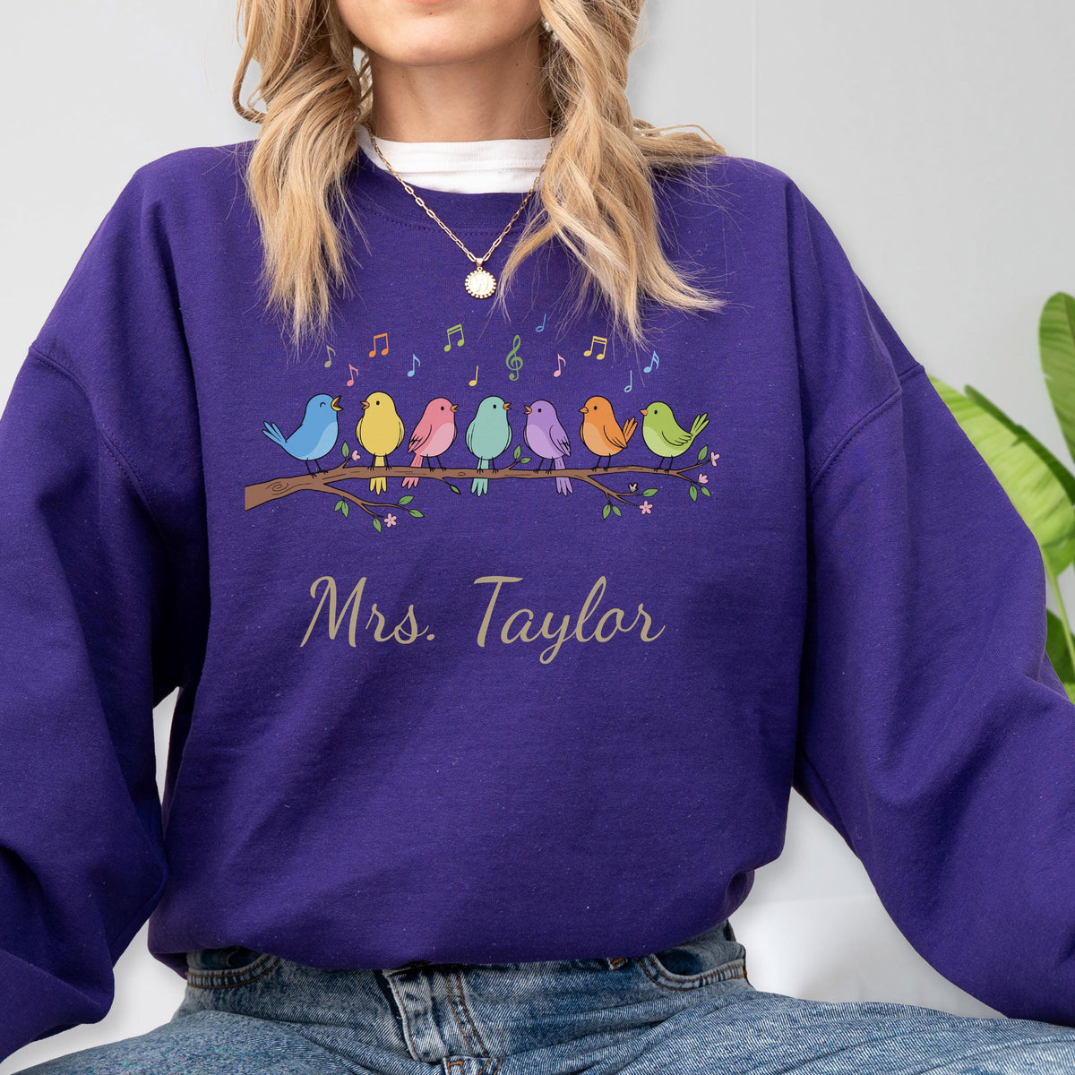 May include: A purple long-sleeve sweatshirt featuring a graphic of six colorful birds perched on a brown branch with musical notes above them. The text "Mrs. Taylor" is printed in a light-colored script font below the bird illustration.
