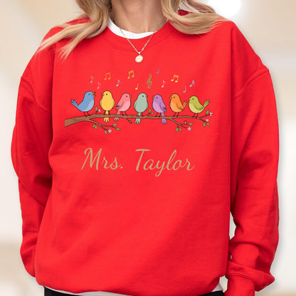 May include: A bright red crewneck sweatshirt featuring a graphic of seven colorful birds perched on a brown tree branch. Above the birds are small musical notes in various colors. The text "Mrs. Taylor" is printed in a gold script font below.