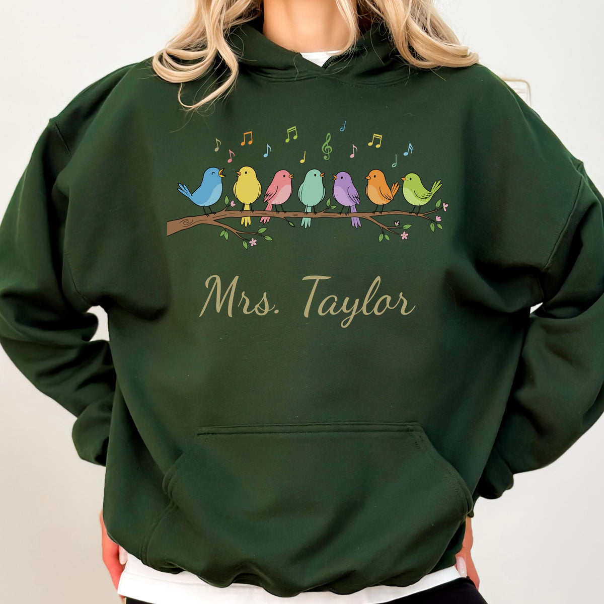 May include: A dark green hooded sweatshirt featuring a graphic of seven colorful birds perched on a brown branch with musical notes above them. Below the birds, the text Mrs. Taylor is printed in a light-colored, cursive script font.