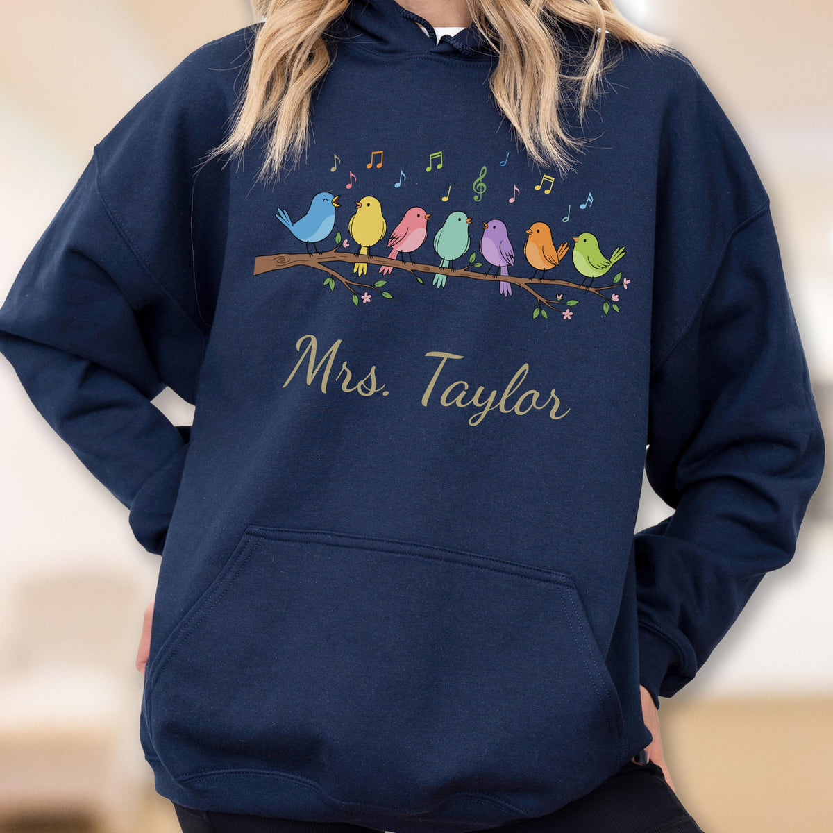 May include: A navy blue hooded sweatshirt featuring a graphic of seven colorful birds perched on a brown branch with musical notes above them. The text "Mrs. Taylor" is printed in a gold script font below the birds on the front of the garment.