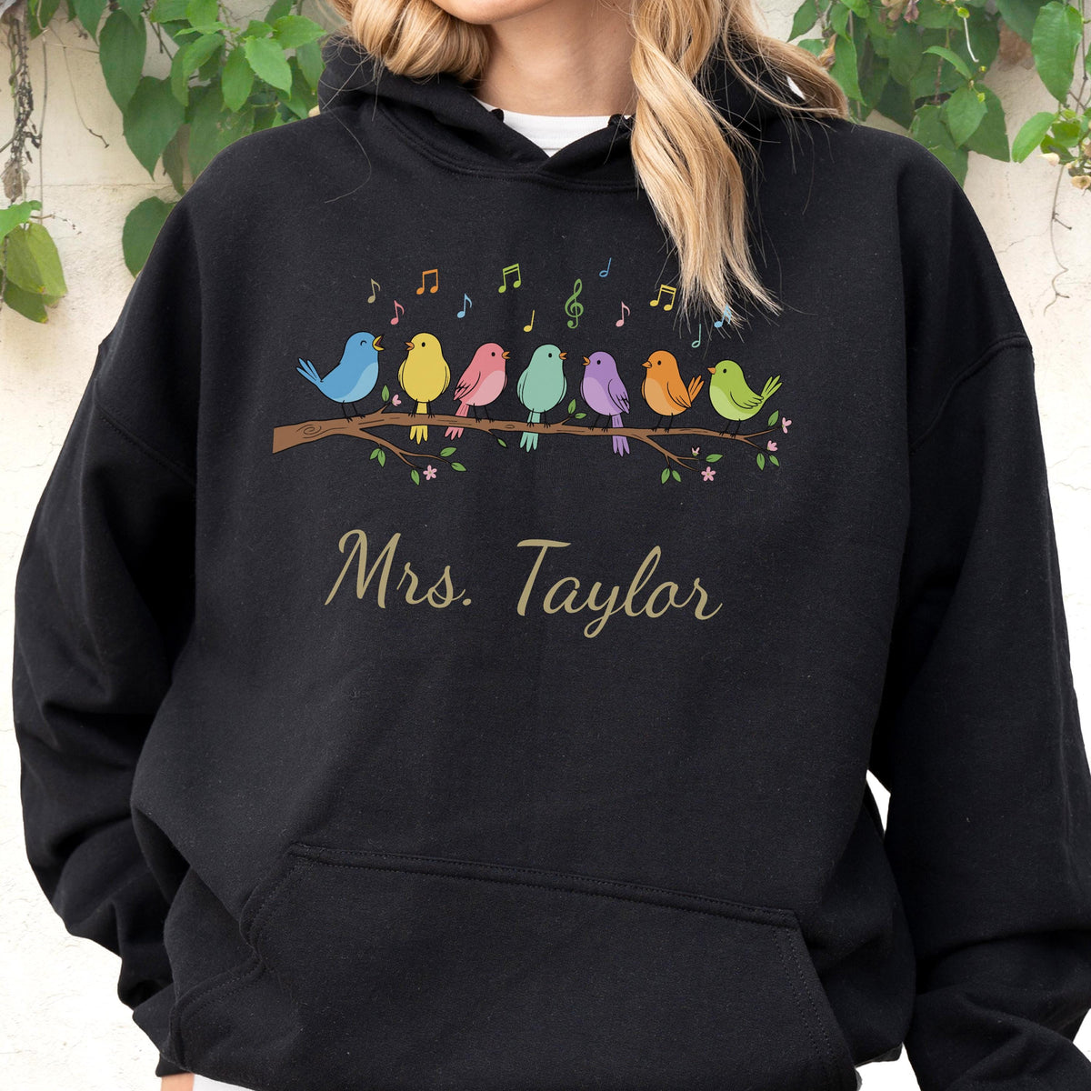 May include: A black hooded sweatshirt featuring a graphic of seven colorful birds perched on a brown branch with musical notes above them. The text "Mrs. Taylor" is printed in a gold script font below the birds on the front of the garment.