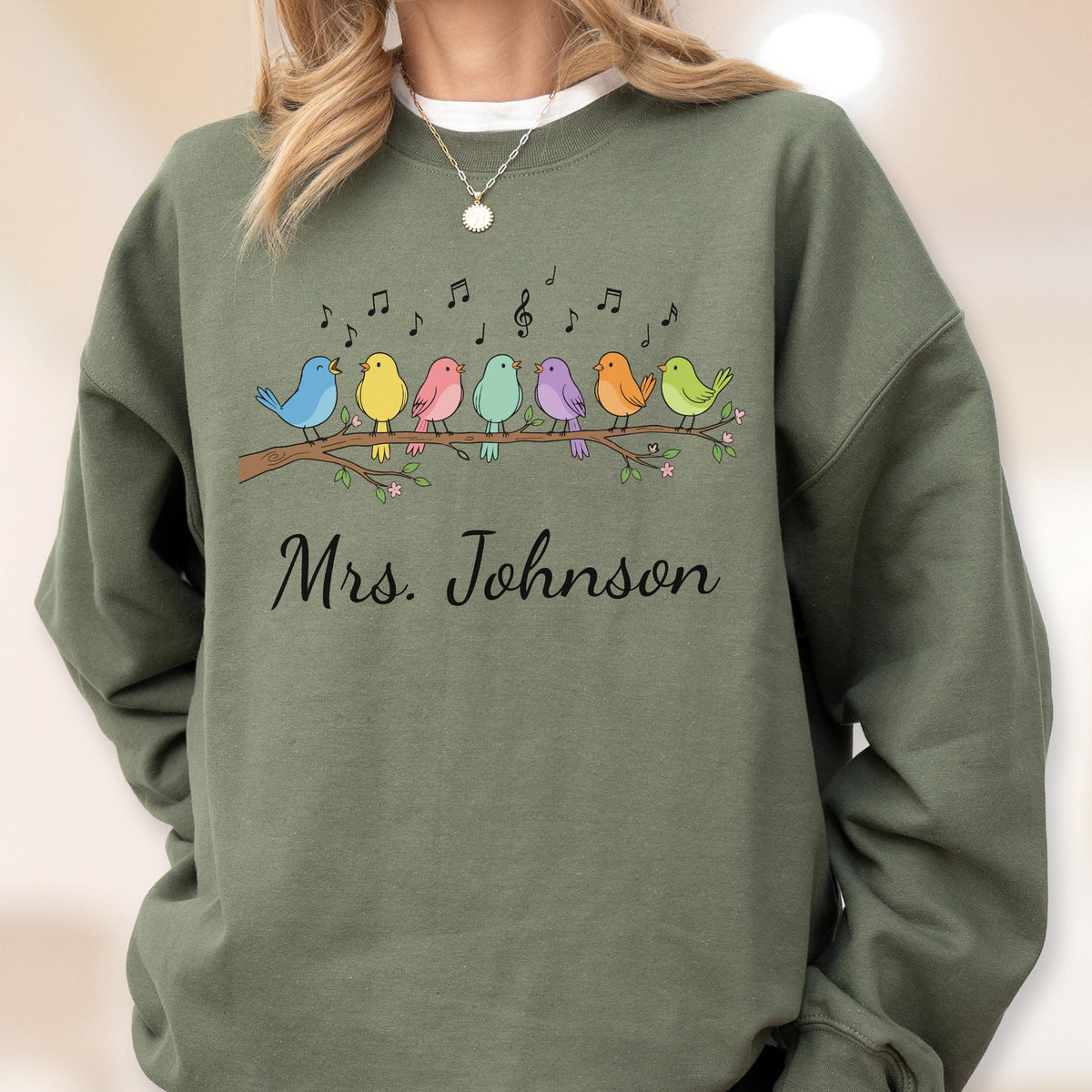 May include: An olive green crewneck sweatshirt featuring a graphic of seven colorful birds perched on a brown branch with musical notes above them. The text "Mrs. Johnson" is printed in black cursive script below the bird illustration.