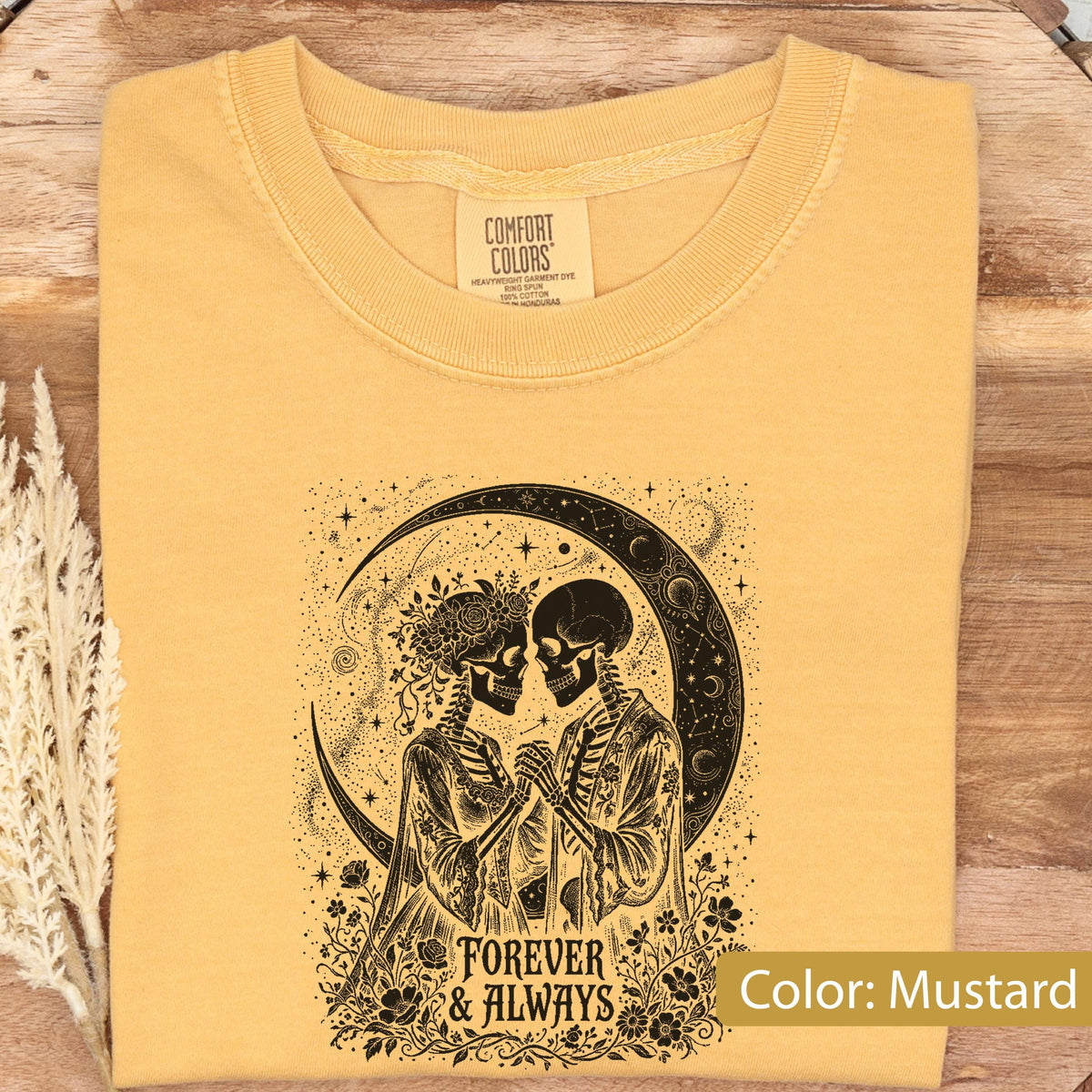 May include: A mustard yellow t-shirt featuring a black graphic print of two skeletons holding hands under a crescent moon. The skeletons are surrounded by flowers and stars. The text "FOREVER & ALWAYS" is printed below the illustration. The shirt has a Comfort Colors brand label at the inner neckline.