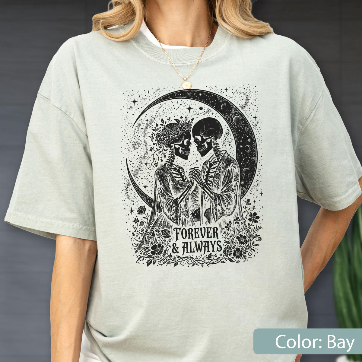 May include: A light sage green t-shirt featuring a black graphic print of two skeletons holding hands under a crescent moon. The design includes floral elements and the text "FOREVER & ALWAYS" centered below the figures in a decorative font.