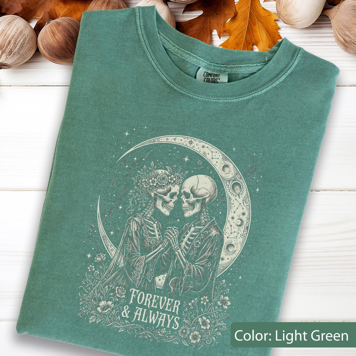 May include: A light green t-shirt featuring a graphic print of two skeletons holding hands in front of a crescent moon. The design includes floral accents and the text "FOREVER & ALWAYS" in a decorative font. The shirt has a Comfort Colors brand label.