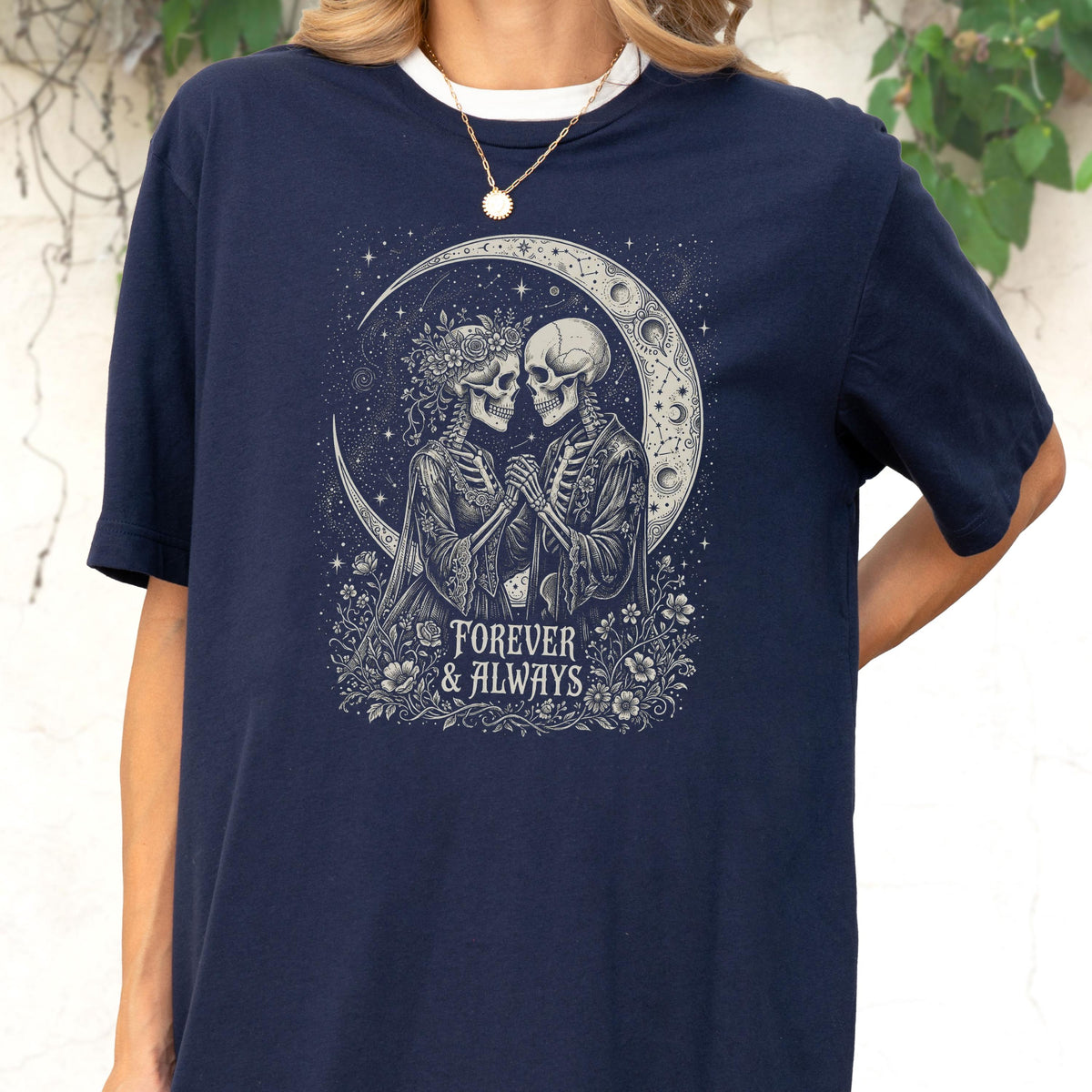 May include: A navy blue t-shirt featuring a graphic print of two skeletons holding hands under a crescent moon. The design includes floral accents and the text "FOREVER & ALWAYS" in a decorative font, set against a starry night sky background.