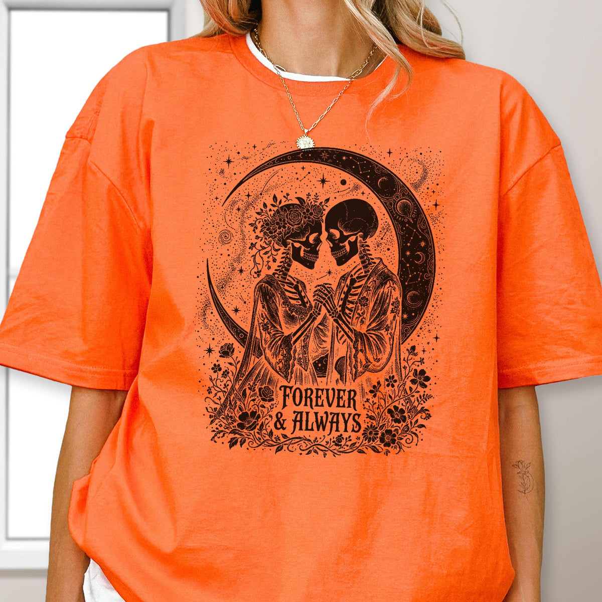 May include: A bright orange short-sleeve t-shirt featuring a black graphic print of two skeletons facing each other, surrounded by flowers and a crescent moon. The text "FOREVER & ALWAYS" is printed in black below the skeletons.