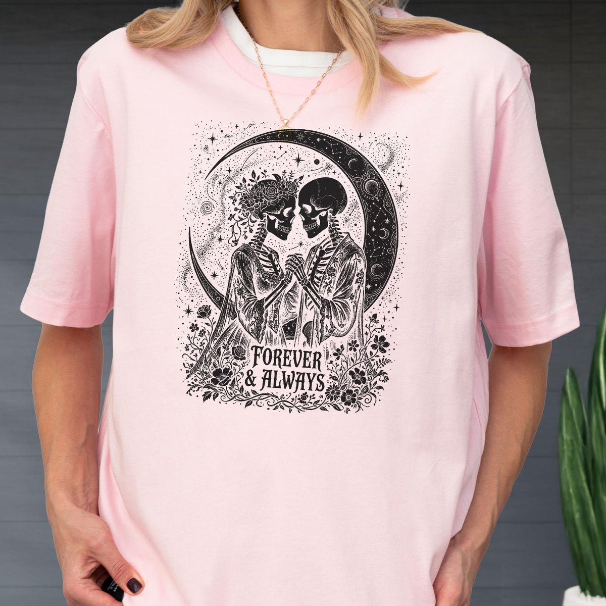 May include: A light pink short-sleeve t-shirt featuring a black graphic print of two skeletons holding hands under a crescent moon. The design includes floral accents and the text "FOREVER & ALWAYS" centered at the bottom of the illustration.