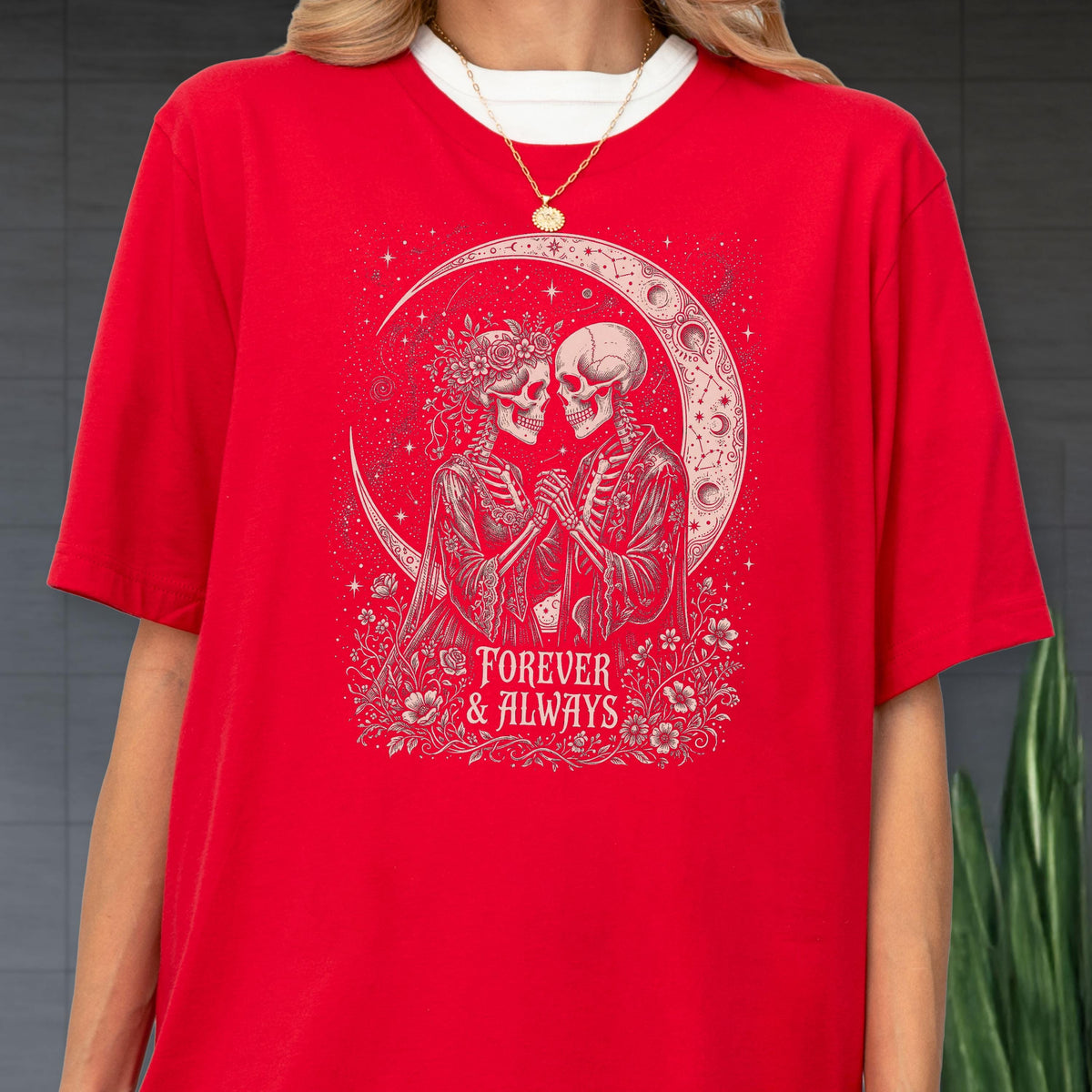 May include: A bright red t-shirt featuring a graphic print of two skeletons holding hands under a crescent moon. The design includes floral accents and the text "FOREVER & ALWAYS" in a decorative font. The shirt has short sleeves and a crew neck.