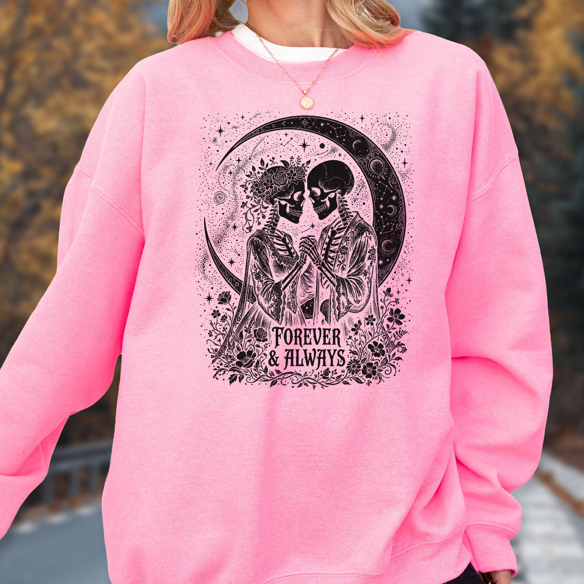 May include: A bright pink crewneck sweatshirt featuring a black graphic print of two skeletons facing each other under a crescent moon. The skeletons are surrounded by floral elements and stars, with the text FOREVER & ALWAYS printed below them.