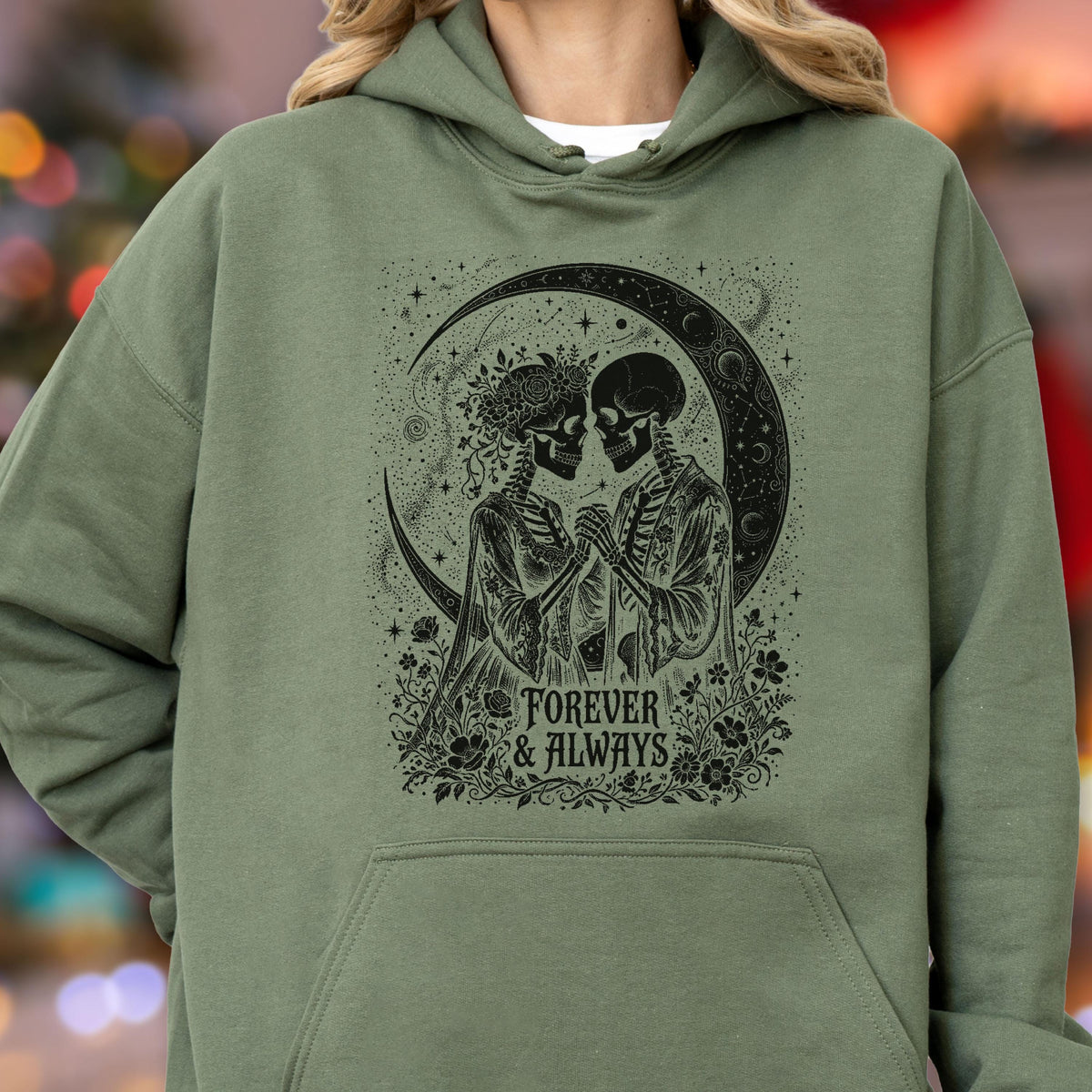 May include: An olive green hooded sweatshirt featuring a black graphic print of two skeletons holding hands under a crescent moon. The design includes floral accents and the text "FOREVER & ALWAYS" centered below the figures in a decorative font.