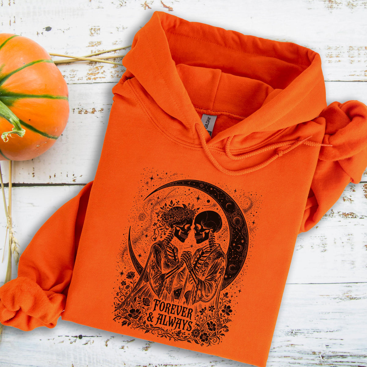 May include: A bright orange hooded sweatshirt featuring a black graphic print of two skeletons facing each other under a crescent moon, surrounded by floral details and the text "FOREVER & ALWAYS" printed in a stylized font on the front.