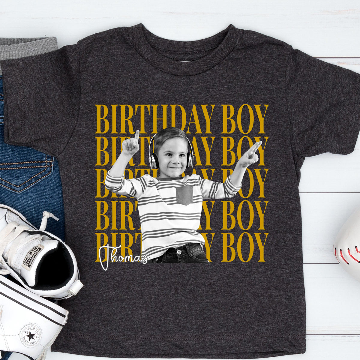 May include: A dark charcoal gray short-sleeve t-shirt featuring a repeating yellow text pattern that reads BIRTHDAY BOY. Centered is a black and white photograph of a child wearing headphones and a striped shirt, with the name Thomas written below.