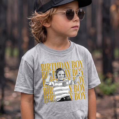 May include: A heather gray t-shirt featuring a repeating mustard yellow text pattern that reads BIRTHDAY BOY. Centered is a black and white graphic of a child wearing headphones and a striped shirt, with the name Thomas written in black script below.
