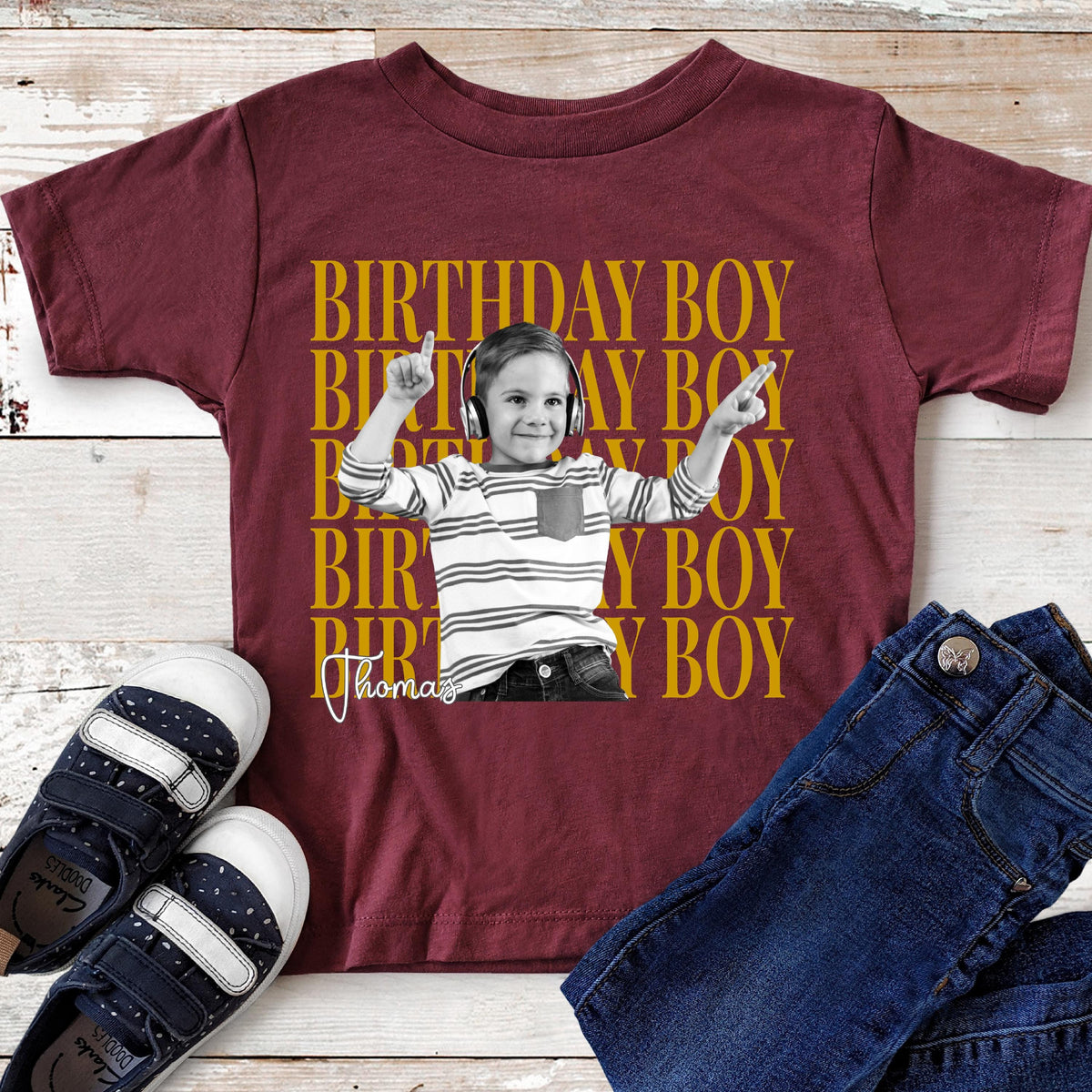 May include: A maroon short-sleeve t-shirt featuring a repeating yellow text pattern that reads BIRTHDAY BOY. Centered is a black and white photograph of a child wearing headphones and a striped shirt, with the name Thomas written in white script.