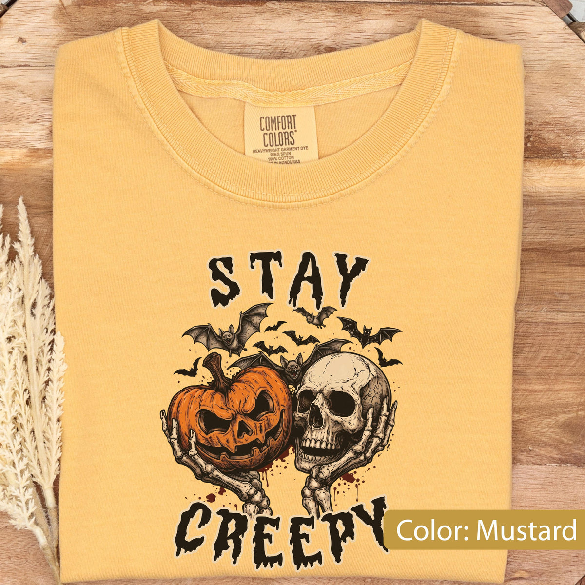 May include: A mustard yellow t-shirt featuring a graphic print of a jack-o'-lantern and a human skull held by skeletal hands, surrounded by flying bats. The text "STAY CREEPY" is printed in black above and below the graphic. The inner neck label reads "COMFORT COLORS".