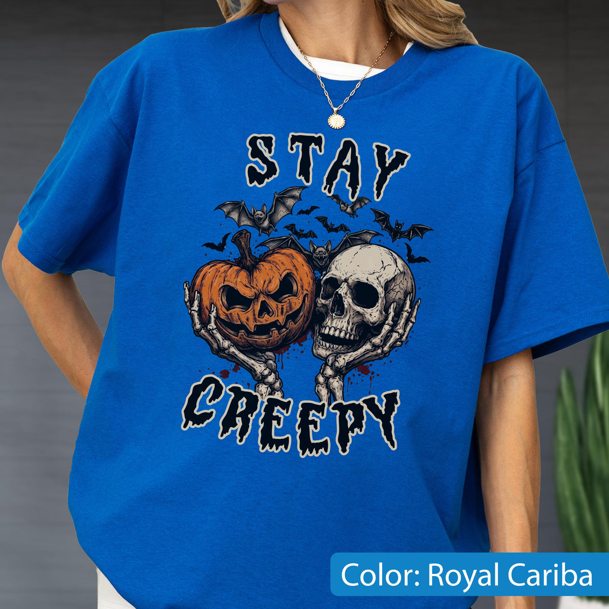 May include: A royal blue t-shirt featuring a graphic print of two skeletal hands holding a jack-o'-lantern and a human skull. Above and below the central illustration, the text reads STAY CREEPY in a black, dripping, stylized font.