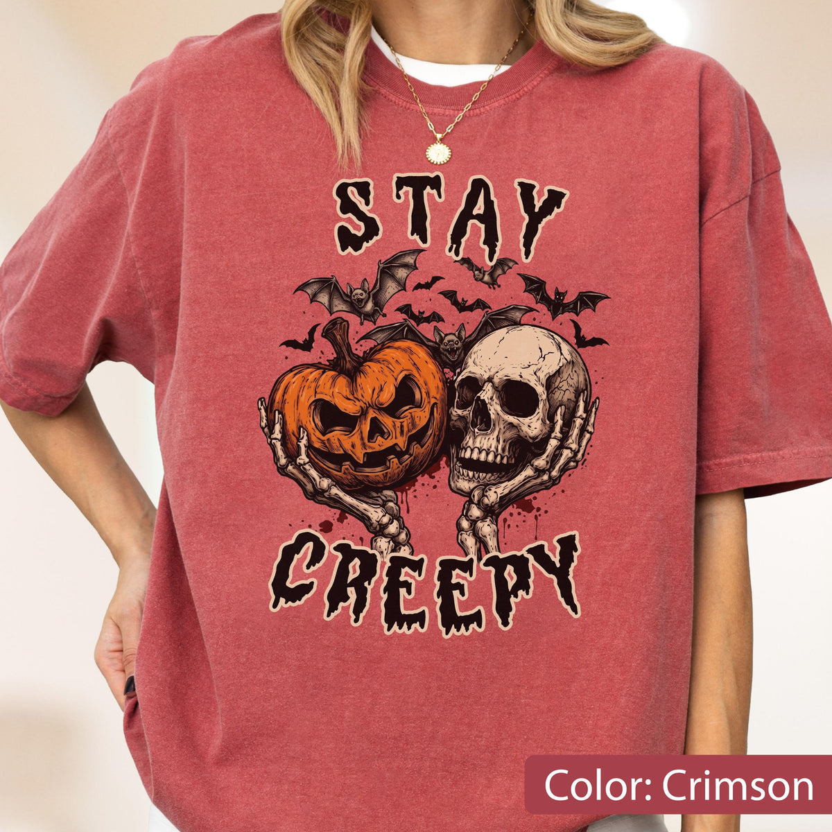 May include: A crimson short-sleeve t-shirt featuring a graphic print of a jack-o'-lantern and a human skull held by skeletal hands. Above and below the illustration, the text reads STAY CREEPY in a jagged black font, surrounded by small bats.
