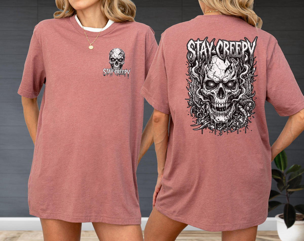 May include: A dusty rose t-shirt featuring a small skull graphic and the text STAY CREEPY on the front left chest, and a large, detailed black and white illustration of a skull surrounded by smoke and vines with the text STAY CREEPY on the back.