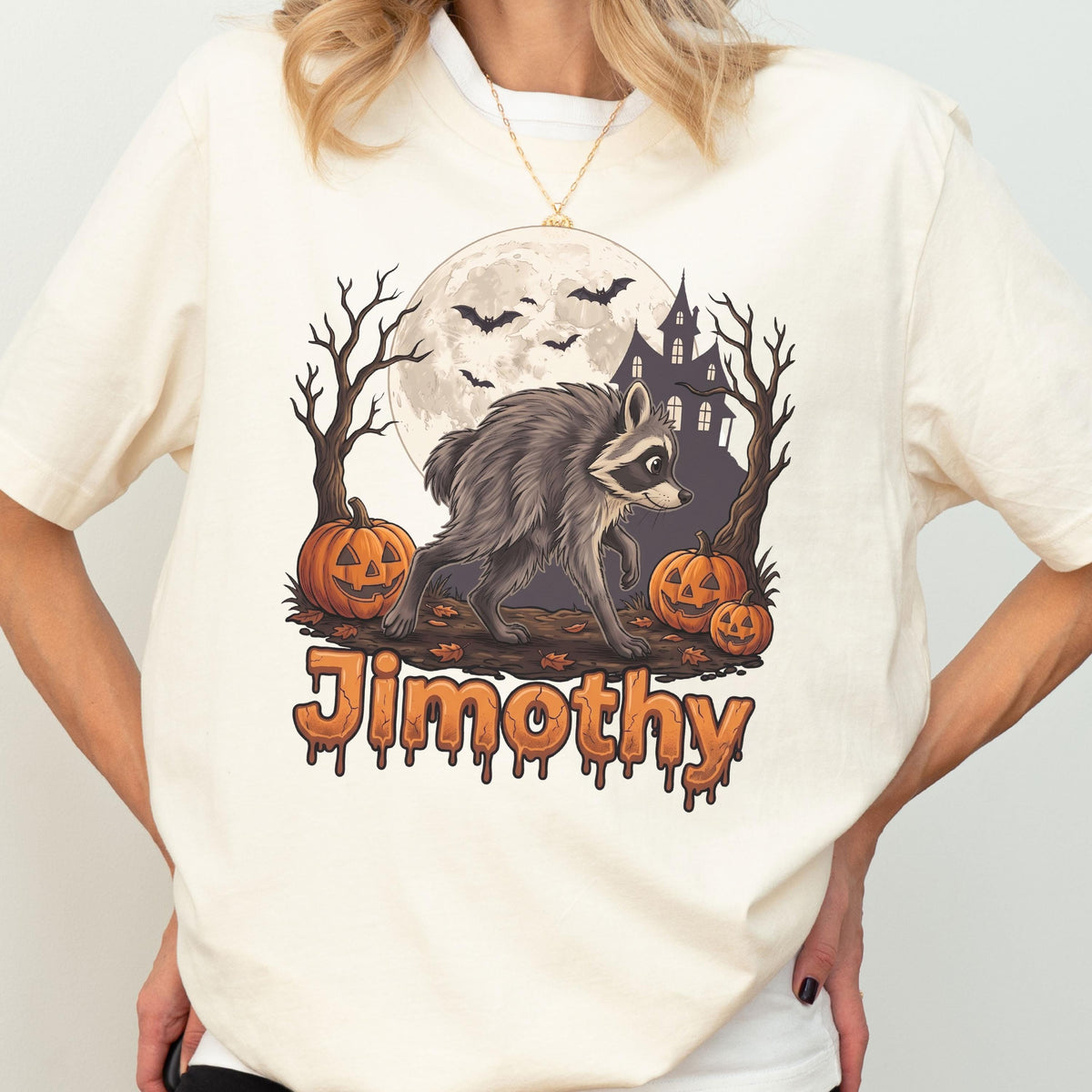 May include: A cream-colored t-shirt featuring a graphic of a raccoon walking in front of a full moon, a haunted house, bare trees, and three jack-o'-lanterns. The name "Jimothy" is printed in a dripping, orange, Halloween-style font below.