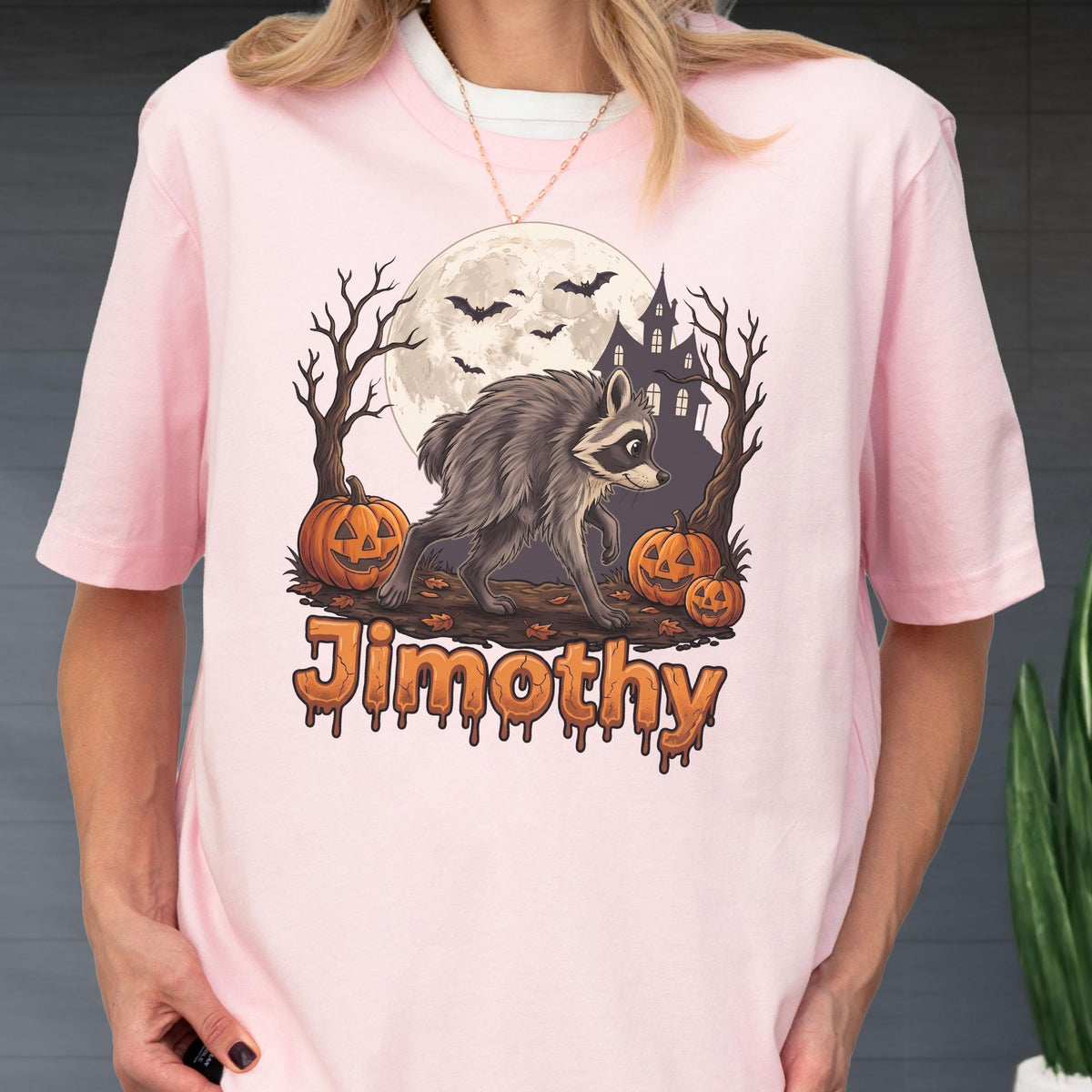 May include: A light pink t-shirt featuring a graphic of a raccoon walking in front of a full moon, a haunted house, bare trees, and three jack-o'-lanterns. The name Jimothy is printed in a dripping orange font at the bottom of the illustration.